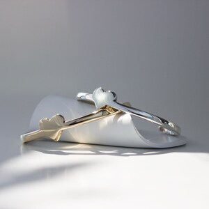 Puede incluir: Dos pulseras en forma de coraz&oacute;n, una dorada y otra plateada, est&aacute;n apiladas sobre una superficie blanca. Las pulseras son simples y elegantes.