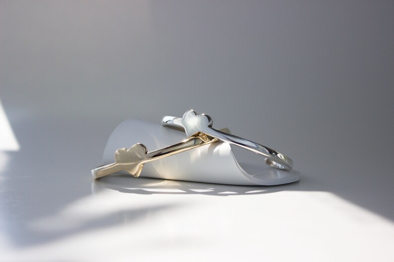 Puede incluir: Dos pulseras en forma de coraz&oacute;n, una dorada y otra plateada, est&aacute;n apiladas sobre una superficie blanca. Las pulseras son simples y elegantes.