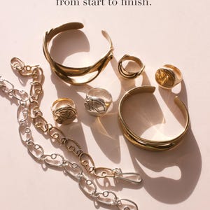 May include: An assortment of gold and silver jewelry, including rings, bracelets, and a chain necklace, is displayed on a light pink surface. The text "All of our jewelry is made by us, from start to finish" and "Handmade in our Portland, Oregon studio" are also visible.