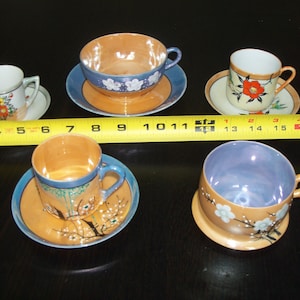 May include: Set of five vintage teacups and saucers with floral designs. The cups and saucers are made of porcelain and have a light blue and orange colour scheme. The floral designs are painted in gold, black, and white.