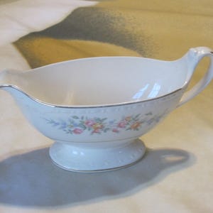May include: A white porcelain gravy boat with a floral design and a gold trim. The boat has a handle and a pedestal base.
