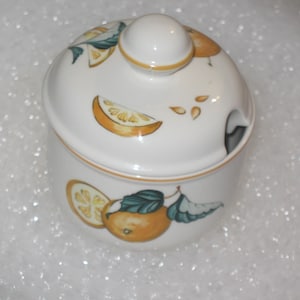 May include: A white ceramic sugar bowl with a lid featuring a round knob and a small notch for a spoon. The exterior is decorated with painted orange fruit, green leaves, and sliced citrus segments in shades of orange and yellow.