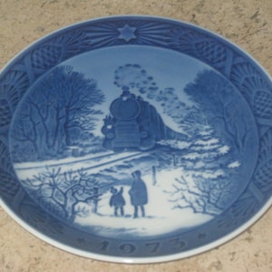 May include: Blue and white porcelain Christmas plate featuring a train with a steam engine and two figures walking in a snowy landscape. The plate has a blue and white border with a star and the year 1975.
