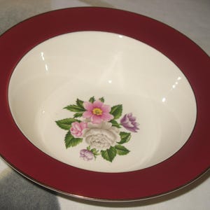May include: A round, white ceramic bowl with a wide, claret-coloured rim and a gold trim. The bowl's interior features a floral design with pink, purple, and white roses and green leaves. A decorative piece for serving.