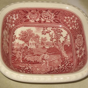 May include: A square ceramic bowl with a cream-colored base and a deep red transferware pattern. The interior features a detailed pastoral scene with figures, trees, and a house, surrounded by a decorative floral border along the rim.