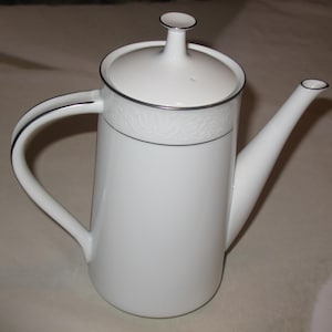 Może przedstawiać: Biały porcelanowy dzbanek do kawy z pokrywką i zakrzywionym uchwytem. Dzbanek ma srebrne obramowanie wokół góry, pokrywki i wylewki. Górna część dzbanka ma dekoracyjny kwiatowy wzór. Dzbanek jest przeznaczony do serwowania kawy.