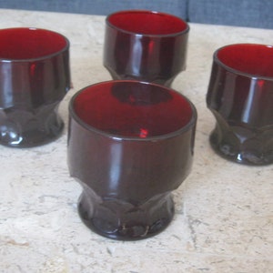 May include: Four dark red glass tumblers with a textured, faceted design. The glasses are empty and sitting on a light-coloured surface.