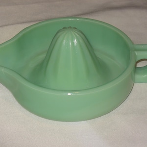 Puede incluir: Un exprimidor de cítricos vintage de vidrio verde jade. El exprimidor presenta un reamer en el centro, un pico vertedor y un asa con un agujero. El diseño es simple y funcional, perfecto para exprimir zumo fresco.