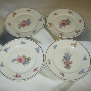 May include: Four cream-coloured ceramic plates with a gold rim and a floral design. The floral design features pink, blue, and yellow flowers. Small floral accents are scattered around the rim.