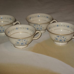 May include: Four white teacups with a delicate blue floral pattern and gold trim. Each cup has a handle and sits on a small footed base. The cups are arranged on a light-colored surface.