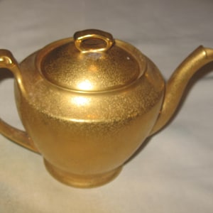 May include: A gold-colored teapot with a matching lid. The teapot has a rounded body, a curved handle, and a spout. The surface has a textured appearance. The lid has a small handle. The teapot is likely for serving tea.