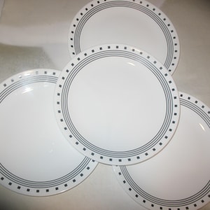 Puede incluir: Un juego de tres platos llanos circulares de color blanco con un patrón decorativo en el borde que consiste en tres finos anillos concéntricos grises y un borde exterior de pequeños cuadrados grises espaciados uniformemente.
