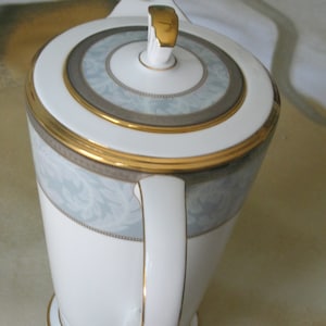 Może przedstawiać: Biały ceramiczny czajnik z pozłacanym wykończeniem i niebiesko-białym wzorem kwiatowym. Czajnik ma pokrywkę z pozłacanym uchwytem.