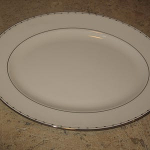 Puede incluir: Una fuente ovalada blanca con una fina línea negra y ribete dorado. El borde presenta un patrón repetitivo de pequeños puntos negros. Este elegante plato es adecuado para servir aperitivos, entrantes o postres.