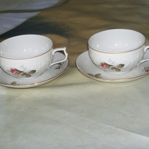 May include: Two white teacups and saucers with a floral design. The cups have a gold rim and the saucers have a gold band around the edge.