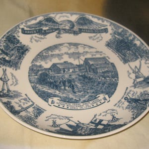 Puede incluir: Un plato blanco con un diseño azul. El plato presenta una escena central de un evento histórico, rodeada de ilustraciones y texto. El texto dice "I HAVEN'T A MAN WHO IS AFRAID TO GO".