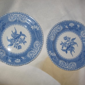 May include: Two blue and white ceramic plates. The plates feature a floral design in the center and a decorative border. The plates are round and have a scalloped edge.