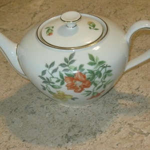 May include: A white teapot with a floral design. The teapot has a lid with a gold trim and a floral design. The teapot is sitting on a light brown surface.
