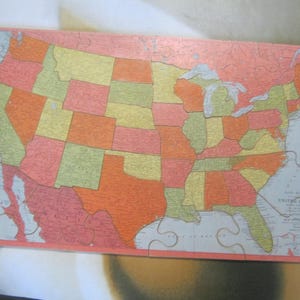 May include: A colorful United States map puzzle with each state in a different color. The puzzle is made of cardboard and has a pink border. The states are in shades of orange, yellow, green, and pink.