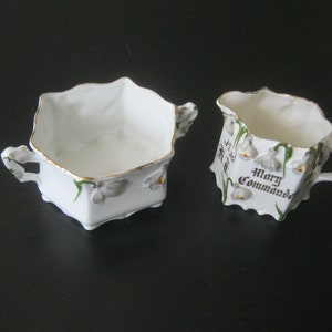 May include: White porcelain sugar bowl and creamer set with floral designs and gold accents. The creamer has the text "Mary Commanda" on the side.
