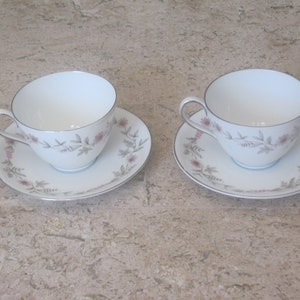 May include: Two white teacups and saucers with a floral pattern in shades of pink and green. The cups have a silver rim and the saucers have a silver band around the edge.