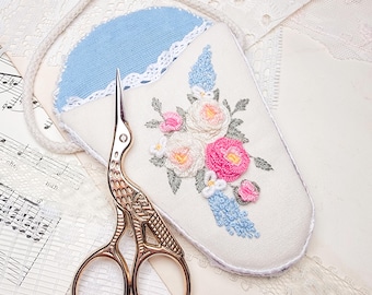 May include: A gold-colored stork-shaped embroidery scissors next to a cream-colored needle case with a blue top and floral embroidery. The needle case has a white lace trim and a white cord. The embroidery features pink, white, and blue flowers.