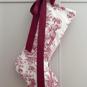 Könnte beinhalten: Ein Weihnachtsstrumpf mit weißem Hintergrund und einem roten Toile-Muster mit Figuren und Blättern. Oben ist eine dunkelrote Schleife gebunden. Der Strumpf hängt an einer weißen Tür.