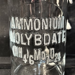 May include: A clear glass bottle with a white label that reads "AMMONIUM MOLYBDATE (NH4)6Mo7O24".