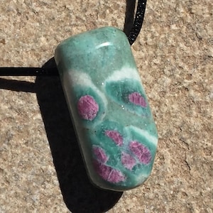 May include: A polished gemstone pendant on a black cord. The pendant is a mix of teal and white with vibrant pink spots. The stone is smooth and rounded, suggesting a natural, organic shape. The cord is thin and black.