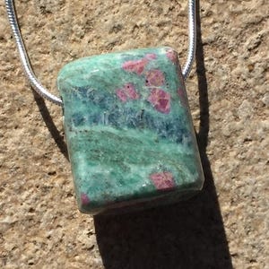 May include: A rectangular pendant featuring a polished stone with a mix of green and blue hues, accented with small pink spots. The stone is strung on a silver chain, creating a unique necklace.