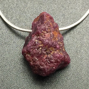 May include: A raw, uncut ruby gemstone pendant on a silver chain. The ruby is a deep red-purple color with a rough, textured surface. The necklace is displayed on a gray surface.
