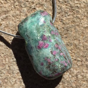 Puede incluir: Un colgante rectangular pulido con una piedra natural de rubí en zoisita. La piedra es una mezcla de tonos verdes y rosas, con una cadena plateada adjunta. El colgante se muestra sobre una superficie texturizada de color marrón claro.