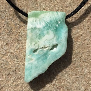 May include: A pendant featuring a polished, irregular slab of light blue and white Larimar stone with natural surface indentations, suspended from a thin black cord. The stone displays a marbled, cloud-like pattern with varying shades of turquoise.