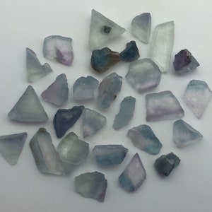May include: A collection of 25 blue and purple fluorite crystals of various shapes and sizes. The crystals are translucent and have a slight shimmer.