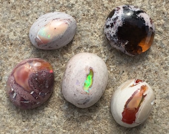 May include: Five polished oval-shaped opal stones with different colours and patterns. The stones are arranged in a circle on a light brown surface.