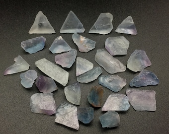 以下が含まれることがあります： 紫、青、透明の様々な色合いの蛍石の結晶20個の集合。結晶は粗く、自然な研磨されていない外観をしています。