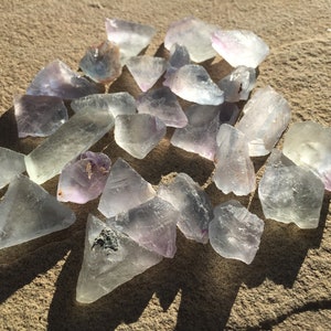 May include: A collection of pale green and purple fluorite crystals, some with a slight iridescent sheen, scattered on a sandy surface.