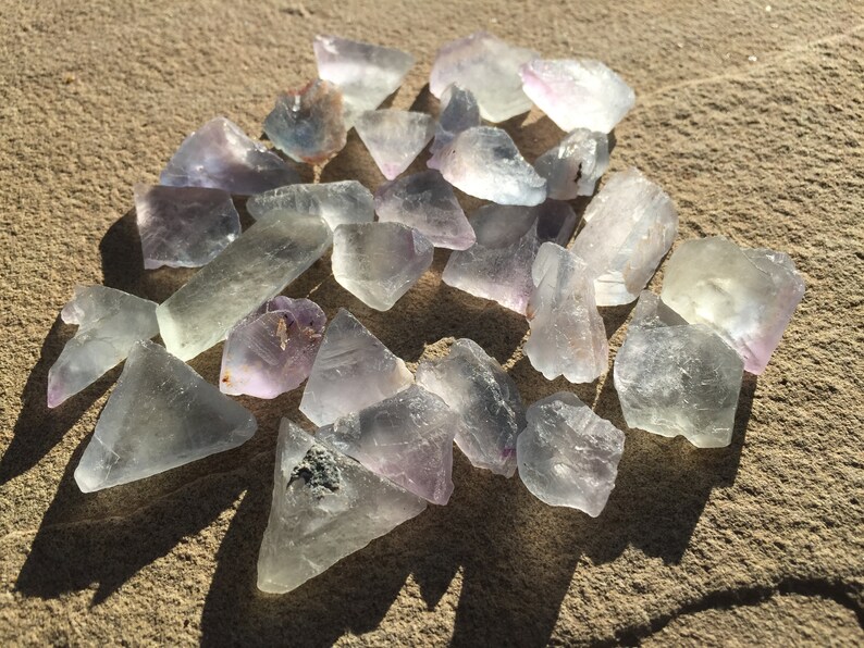 May include: A collection of pale green and purple fluorite crystals, some with a slight iridescent sheen, scattered on a sandy surface.