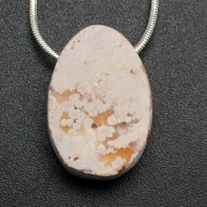 May include: Oval pendant with a cream-colored stone, featuring orange and brown patterns. The stone is suspended from a silver-colored chain. The pendant is set against a dark gray textured background.