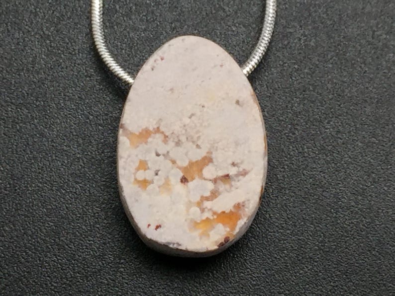 May include: Oval pendant with a cream-colored stone, featuring orange and brown patterns. The stone is suspended from a silver-colored chain. The pendant is set against a dark gray textured background.