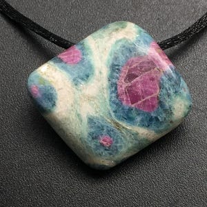 Peut inclure: Un pendentif de forme carrée composé d'une pierre précieuse polie présentant un motif marbré de magenta vif, de bleu sarcelle et de blanc. La pierre est suspendue à un cordon noir fin, révélant une surface lisse et arrondie aux nuances variées.