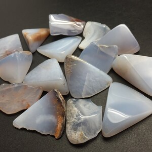 May include: A collection of blue chalcedony tumbled stones, each with a unique shape and pattern. The stones are smooth and polished, with a light blue colour and some brown markings.