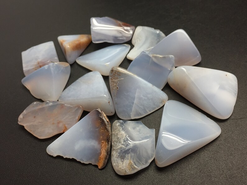 May include: A collection of blue chalcedony tumbled stones, each with a unique shape and pattern. The stones are smooth and polished, with a light blue colour and some brown markings.