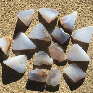 May include: A collection of 14 blue agate stones, some with brown markings, in various shapes and sizes. The stones are arranged on a light brown surface.