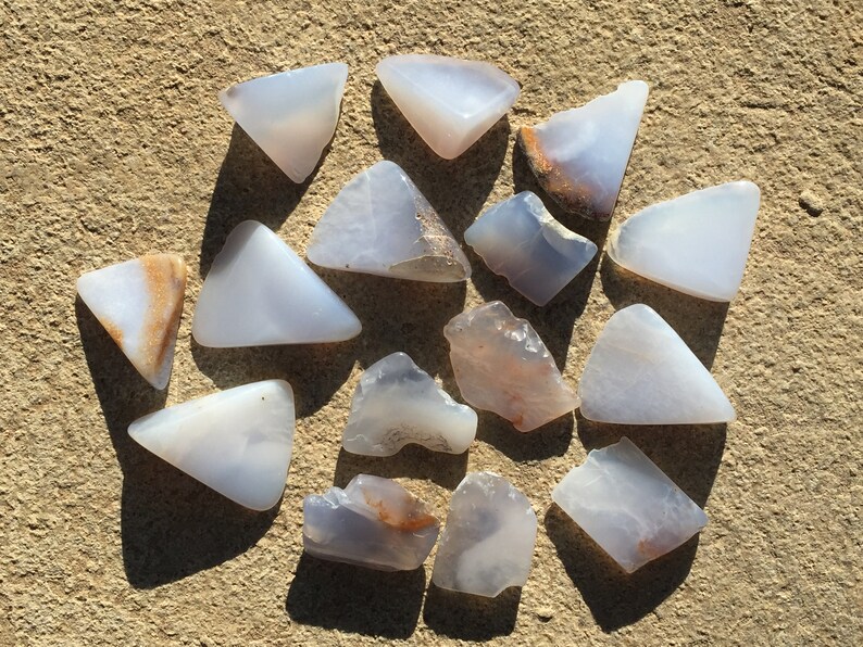 May include: A collection of 14 blue agate stones, some with brown markings, in various shapes and sizes. The stones are arranged on a light brown surface.