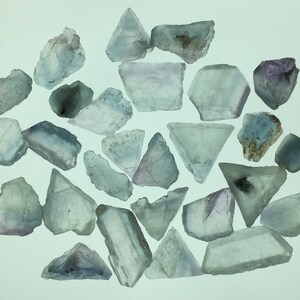May include: A collection of 25 light blue fluorite crystals with some purple and white hues. The crystals are various shapes and sizes, some are triangular, some are rectangular, and some are irregular.