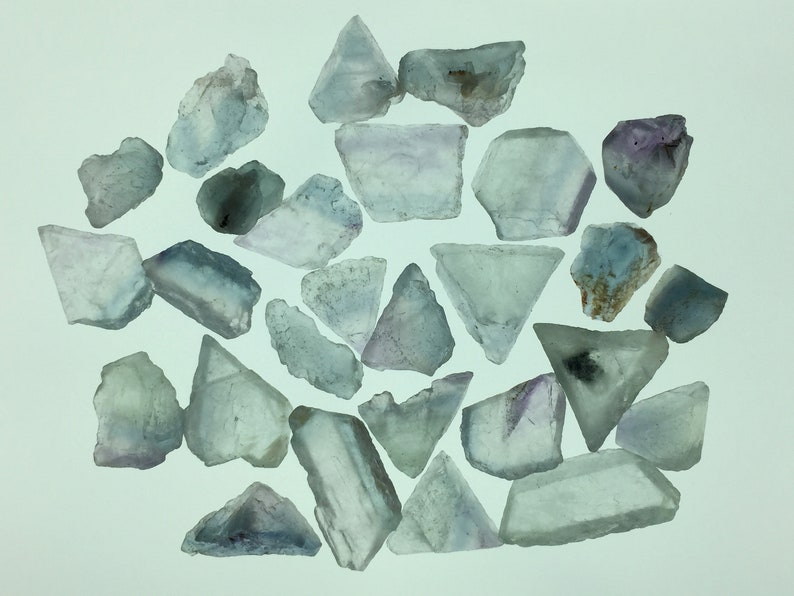 May include: A collection of 25 light blue fluorite crystals with some purple and white hues. The crystals are various shapes and sizes, some are triangular, some are rectangular, and some are irregular.