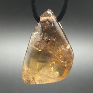 May include: A polished, irregular, translucent amber-colored gemstone pendant suspended from a thin black cord. The stone features internal inclusions, varying shades of yellow and brown, and a smooth, glossy surface texture throughout.