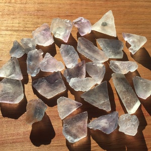 May include: A collection of 25 light purple and clear fluorite crystals, some with a slight green hue, on a wooden surface.