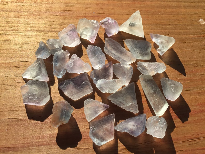 May include: A collection of 25 light purple and clear fluorite crystals, some with a slight green hue, on a wooden surface.